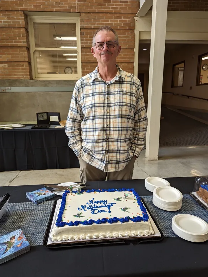 Don Powers standing behind a hummingbird-decorated ‘Happy Retirement Don!’ cake